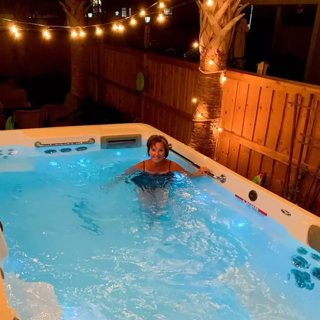 Woman lounging in swim spa in backyard with fence and lights on tree