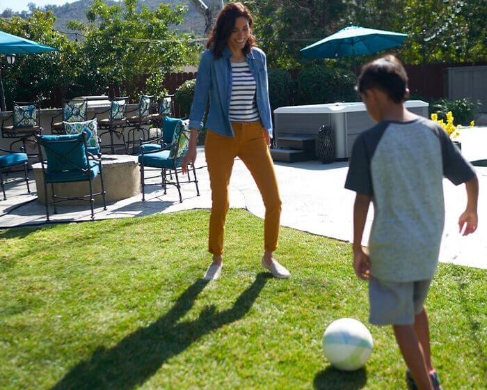 Mom and son playing soccer in the grass in their backayard next to a hot tub and patio furniture